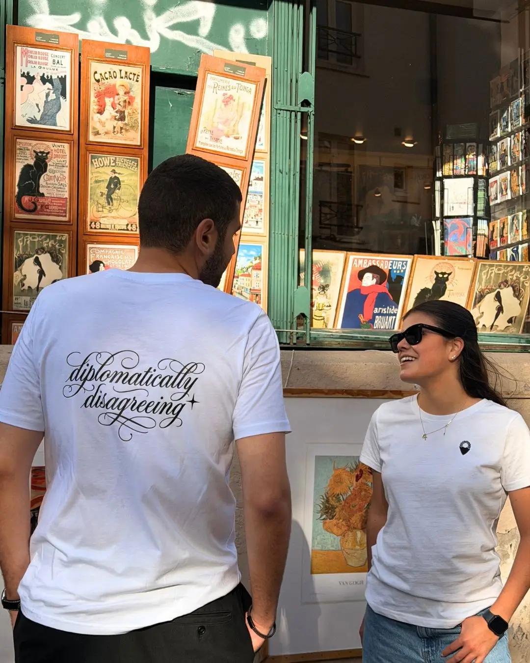 Two people wearing the white mymun t-shirt outside a Paris print shop: one shows "diplomatically disagreeing" in black script on the back, the other the small mymun pin on the front